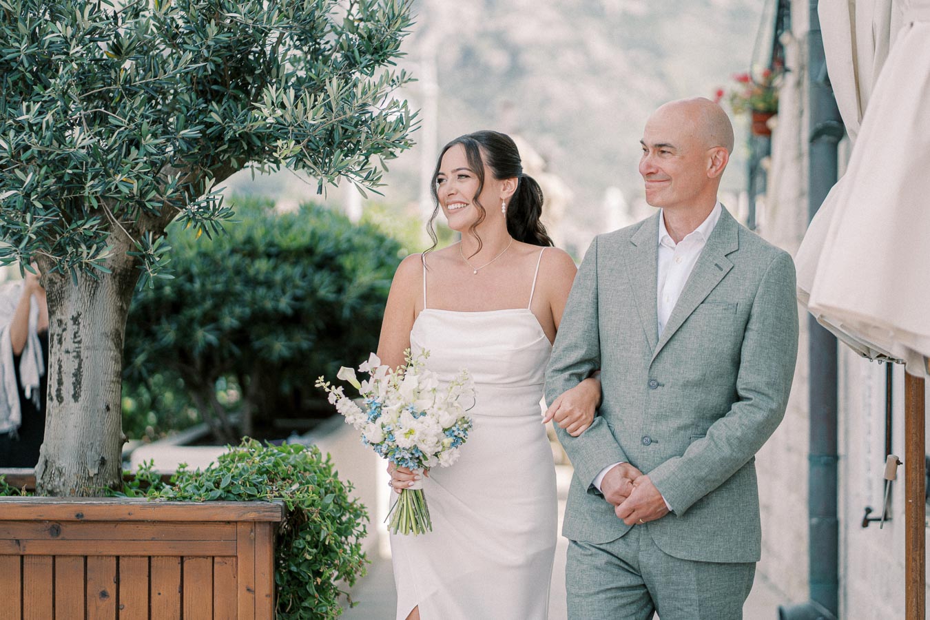 Bride in elegant white dress walking with a man in a gray suit, holding a bouquet of white and blue flowers, under a lush green tree in a serene outdoor setting.