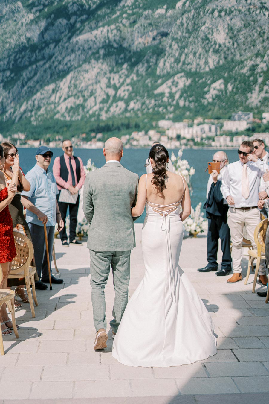 A bride in a white gown and groom in a light grey suit walk down an outdoor aisle, surrounded by seated guests against a backdrop of stunning mountain views and a serene lake.