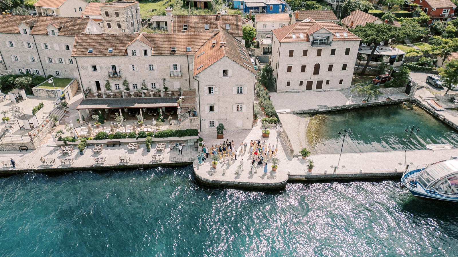 Aerial view of a picturesque coastal village featuring stone buildings with terracotta roofs, a waterfront promenade, and a group of people gathering on a stone platform near the clear blue sea, captured on a sunny day.