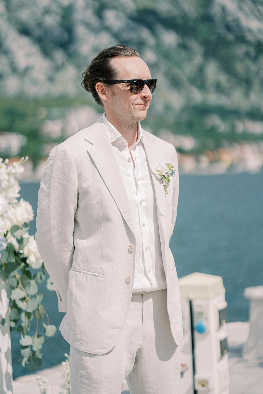Man in a light suit with sunglasses stands by a scenic waterfront, with a mountain landscape and floral decorations in the background, exuding a stylish and relaxed atmosphere.