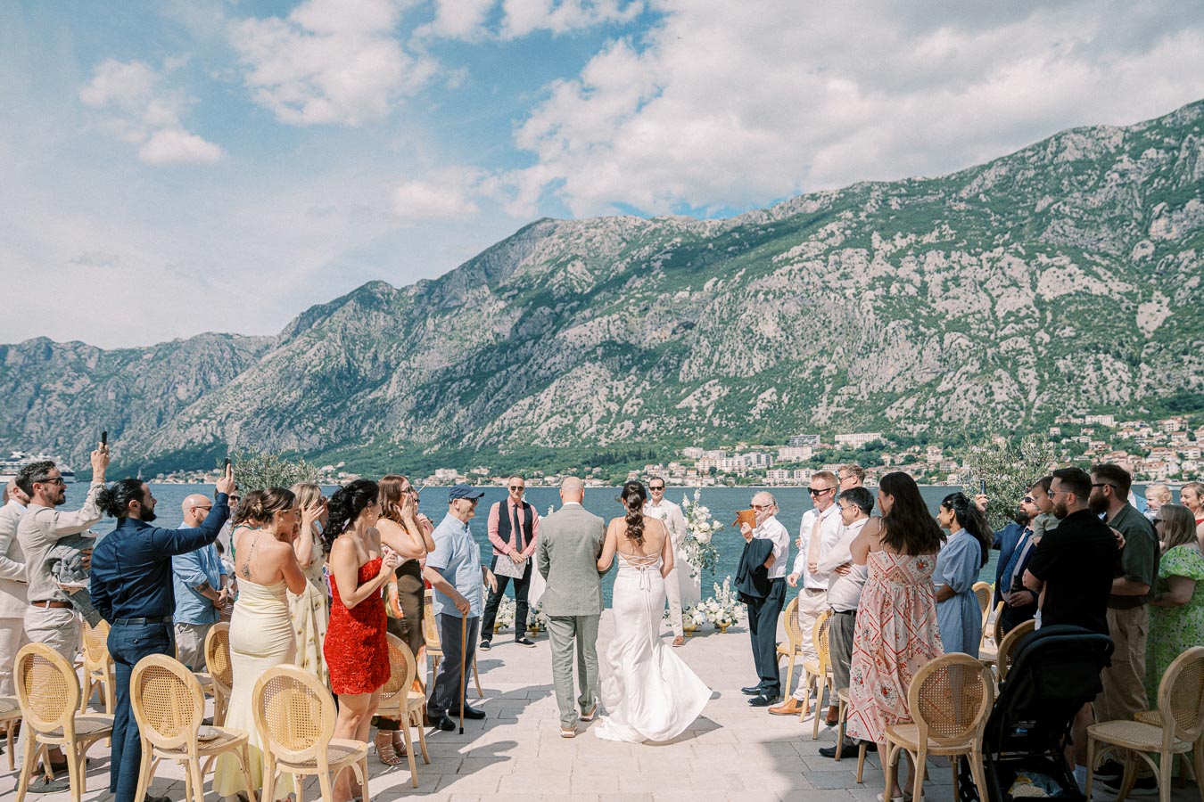 Beachfront wedding ceremony with guests, overlooking mountains and ocean, under a clear blue sky.
