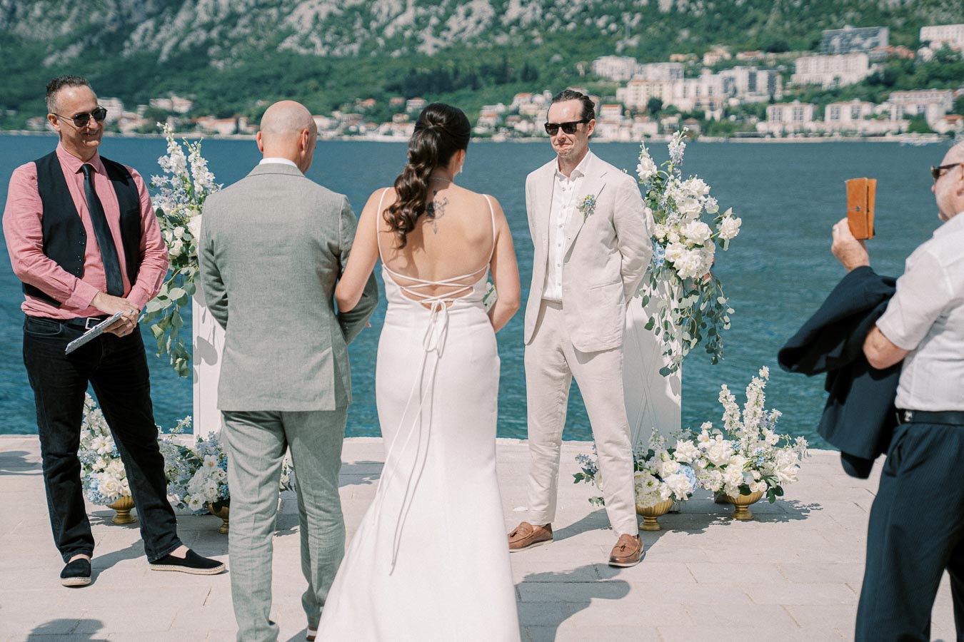 Outdoor wedding ceremony by a scenic lake with a bride in a white dress and groom in light suit, surrounded by floral decorations, with an officiant and guests present.