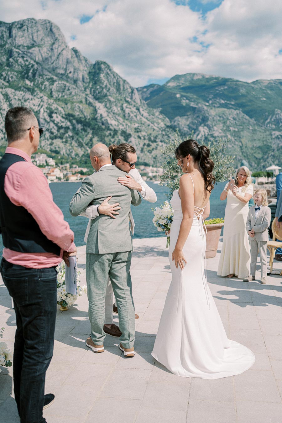 Outdoor lakeside wedding ceremony with guests witnessing a heartfelt embrace between two men, while the bride in a white gown stands nearby against a backdrop of stunning mountains under a blue sky.