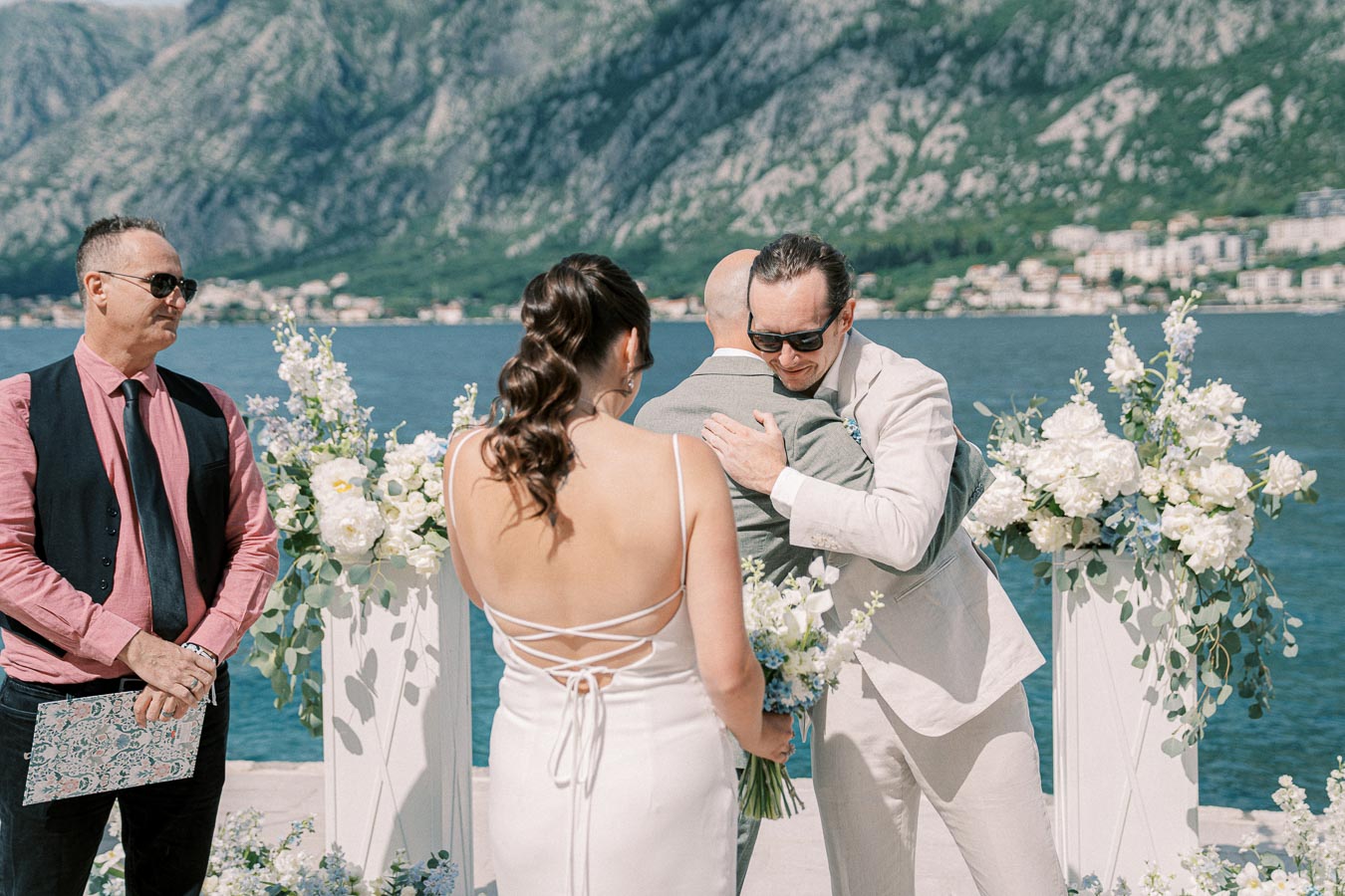 Outdoor wedding ceremony by the sea with mountains in the background, featuring a bride in a white dress holding a bouquet, two men embracing in suits, and a man officiating with floral arrangements nearby.