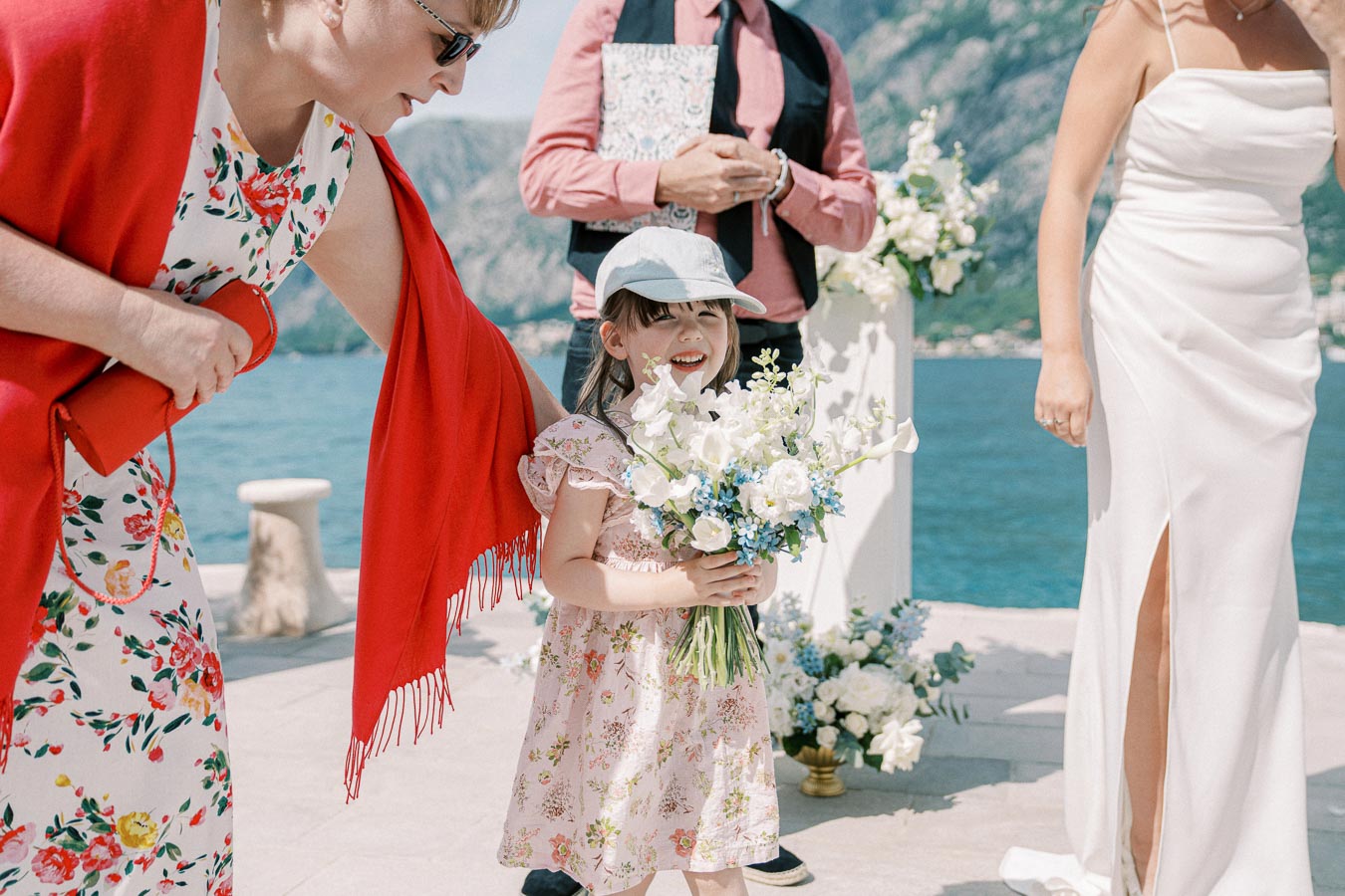 A young girl in a floral dress and hat holding a bouquet of white and blue flowers at an outdoor wedding ceremony by the sea, surrounded by elegantly dressed adults and floral arrangements.