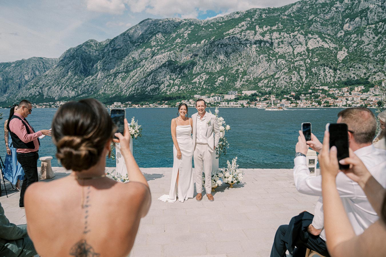 Scenic outdoor wedding ceremony by the sea with a happy couple posing for photos, surrounded by guests capturing the moment against a backdrop of mountains and a coastal town.