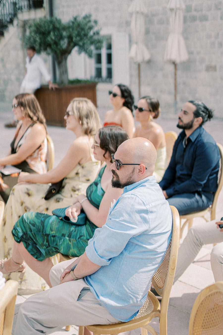 A group of people seated outdoors at an event, wearing summer attire and sunglasses, with a stone building and potted tree in the background.