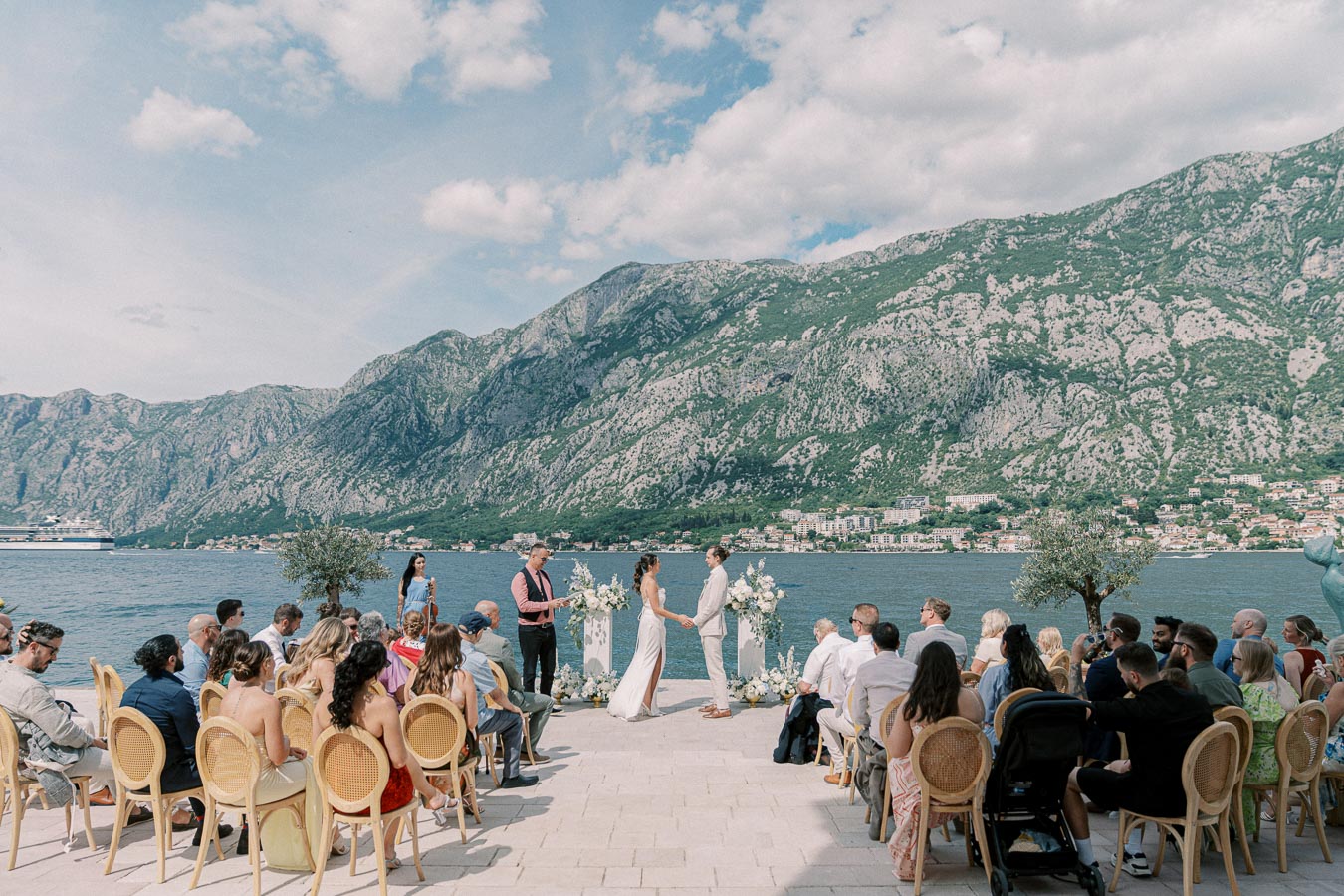 Outdoor wedding ceremony by a scenic lake and mountainous backdrop, featuring a couple exchanging vows surrounded by guests seated on elegant chairs.
