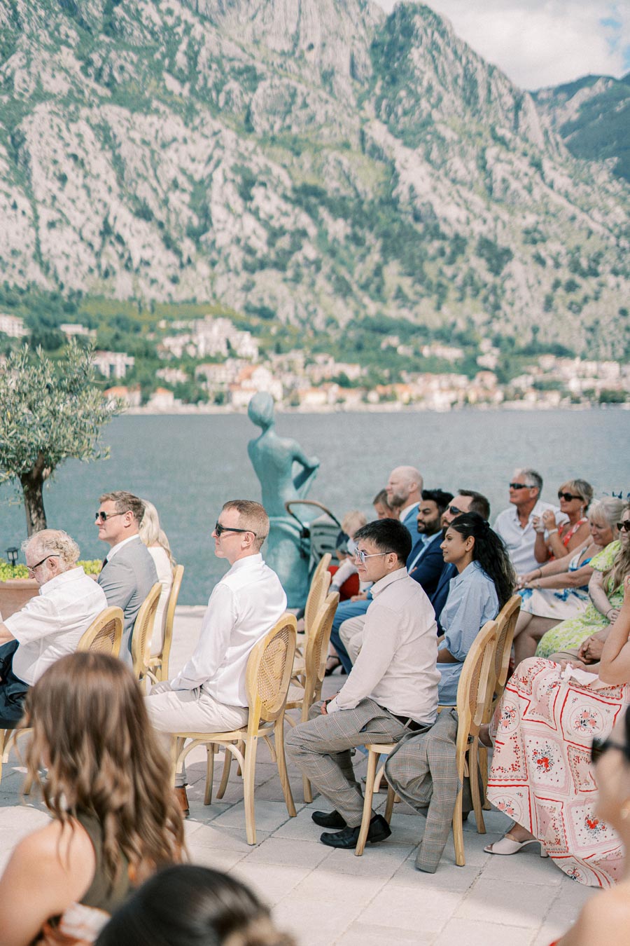 Outdoor wedding ceremony by a lake with guests seated on wooden chairs, scenic mountains in the background, and a sculpture near the water.