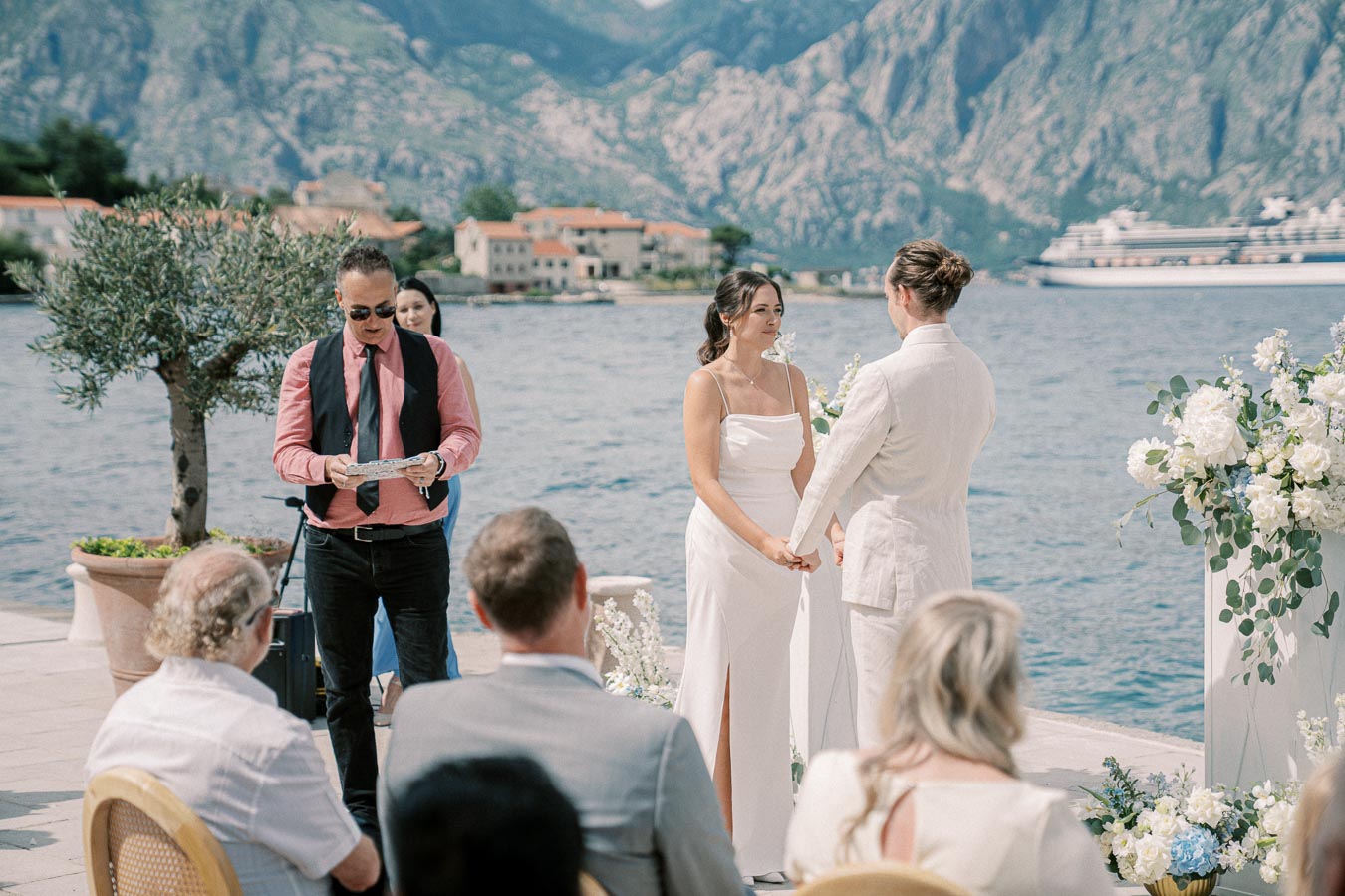 A picturesque outdoor wedding ceremony by the sea, featuring a couple holding hands surrounded by guests and a scenic mountain and water backdrop.
