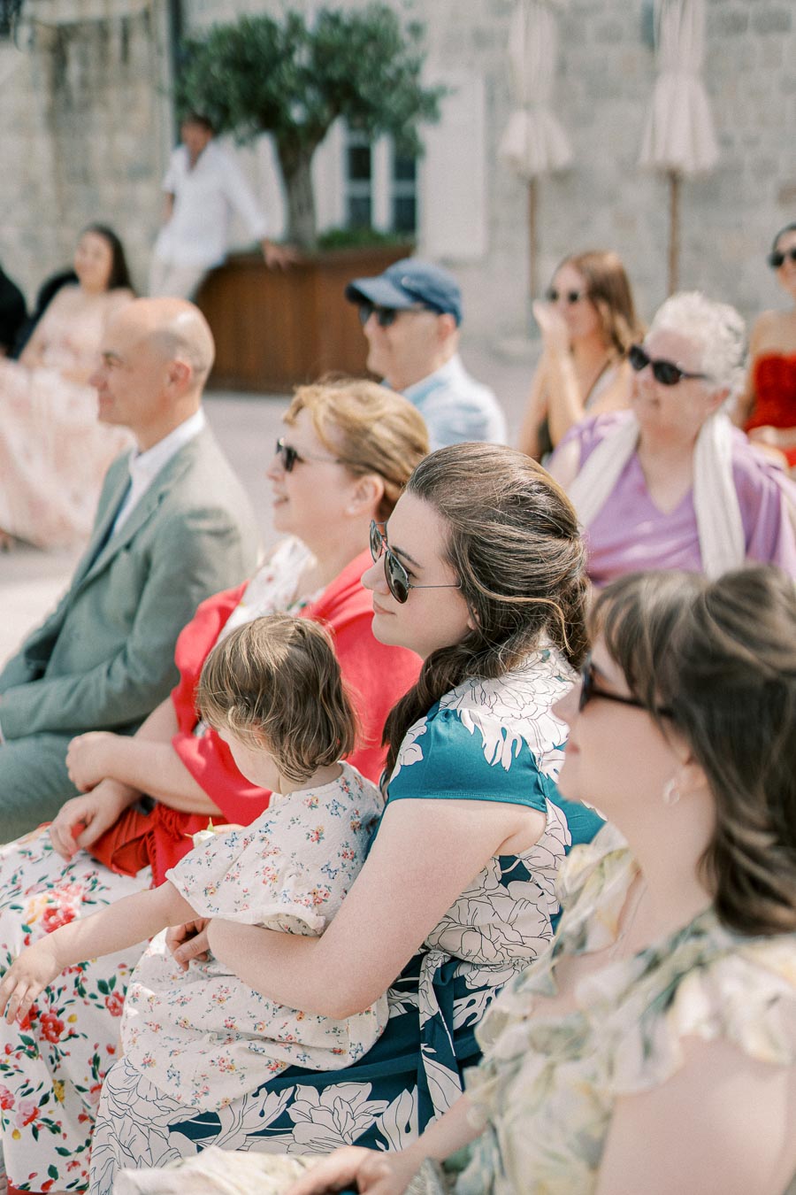A group of people seated outdoors at a daytime event, wearing sunglasses and colorful clothing, with a brick building and plants in the background.