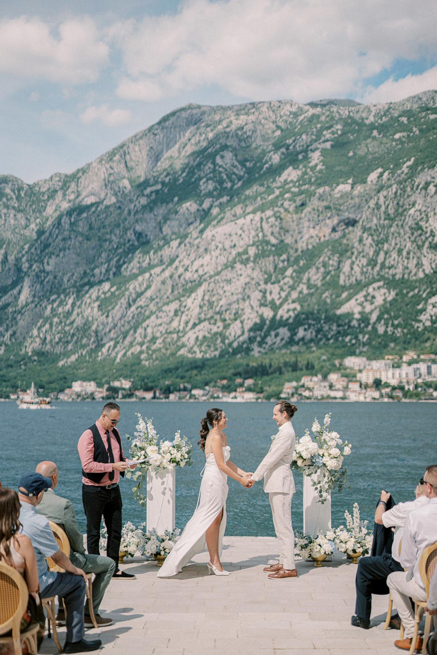 A couple exchanging vows in a scenic outdoor wedding ceremony by a lake, with a mountainous backdrop on a sunny day, surrounded by guests and floral arrangements.