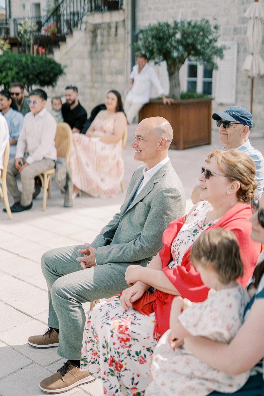 Guests seated outdoors at a wedding ceremony, enjoying the event in formal attire with a backdrop of rustic stone architecture and greenery.