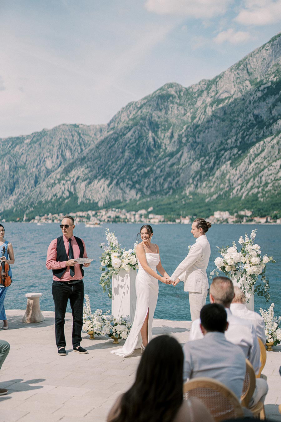 Outdoor waterfront wedding ceremony with a couple exchanging vows surrounded by floral arrangements, set against a backdrop of scenic mountains and a clear blue sky.