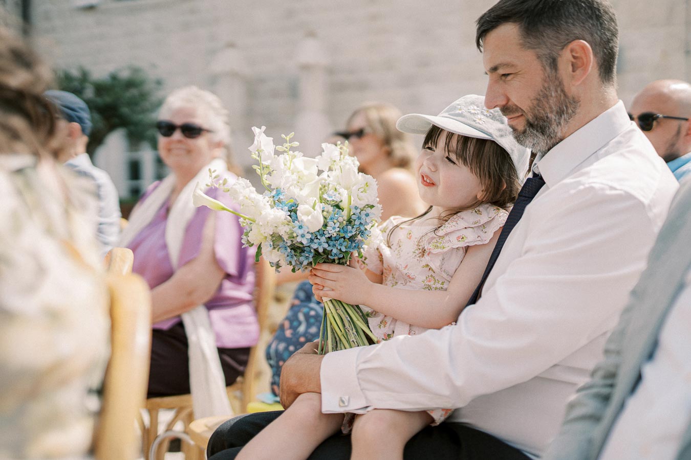 Father holding daughter with bouquet during outdoor event, surrounded by seated guests dressed in summer attire.