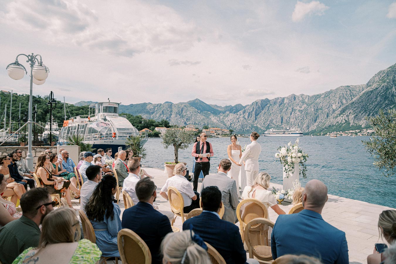 Outdoor waterfront wedding ceremony with a bride and groom exchanging vows, surrounded by guests, against a backdrop of mountains and a docked boat.