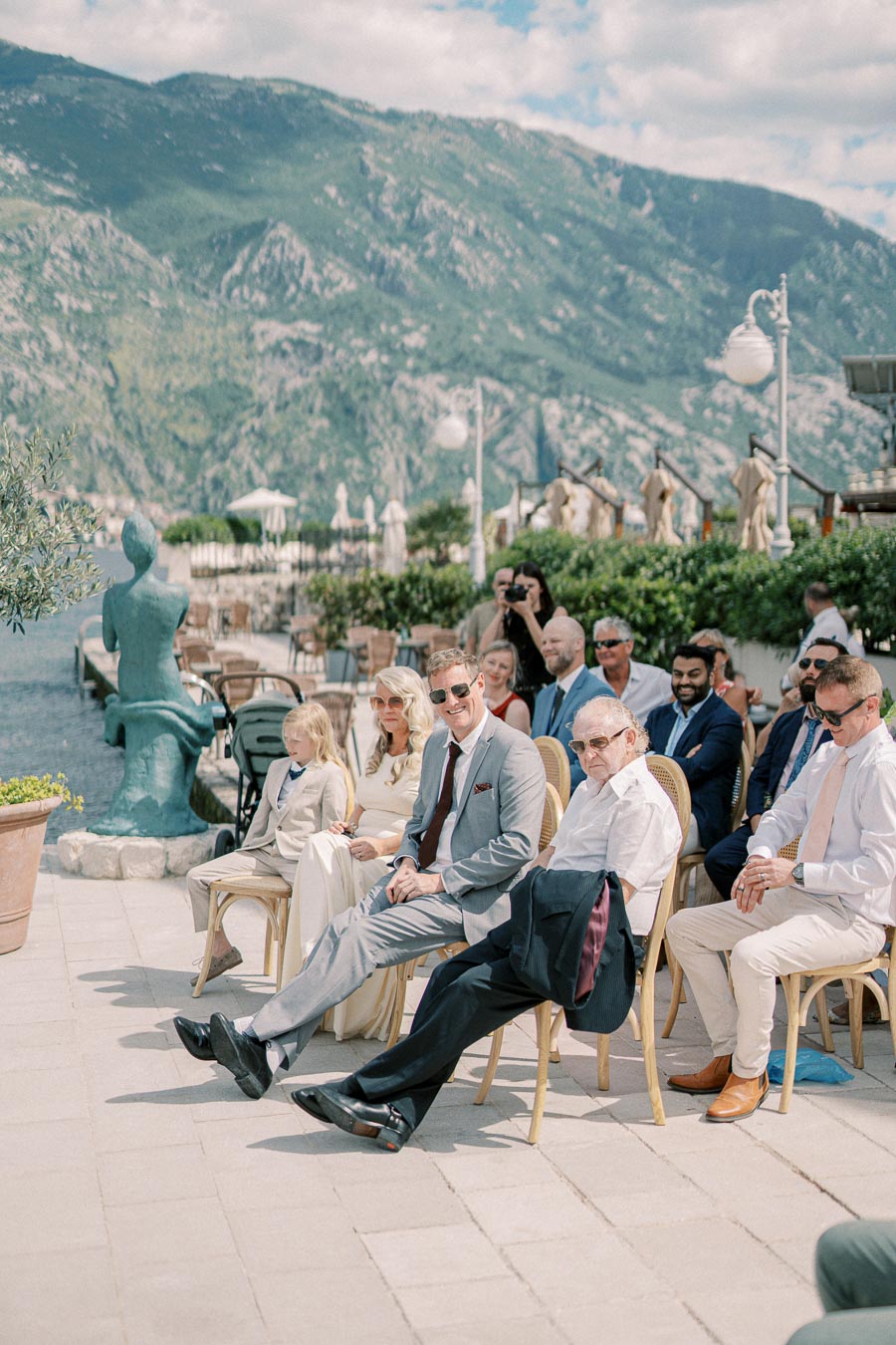 A group of elegantly dressed people seated outdoors at a waterfront event, with a scenic mountainous backdrop and a green statue nearby.