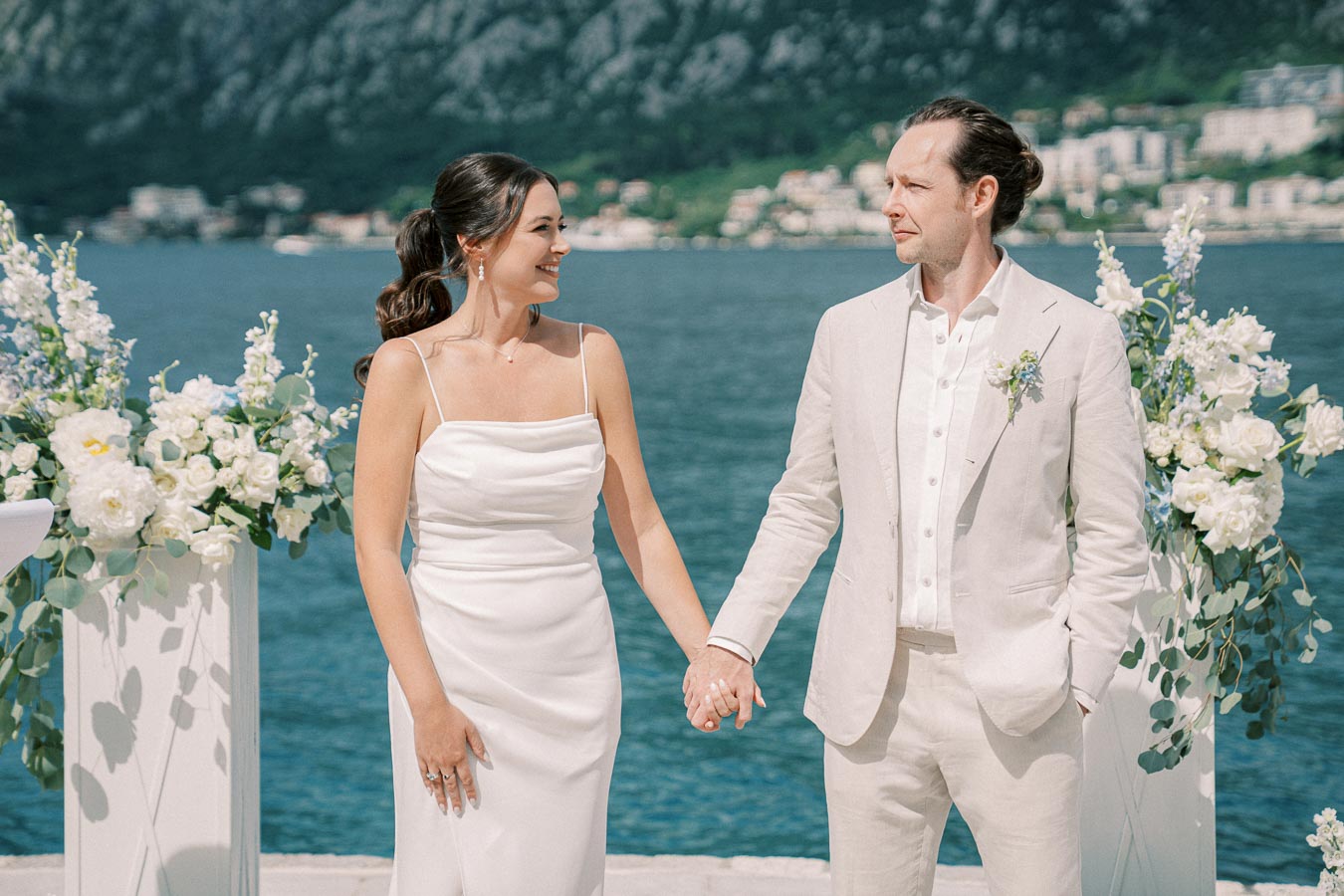 A couple in elegant white attire holds hands at a waterfront wedding ceremony, surrounded by white and blue floral arrangements, with a picturesque mountain and ocean backdrop.