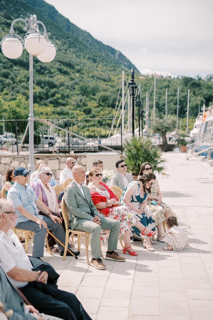 A group of people seated outdoors in a picturesque mountainous area, enjoying a sunny day with yachts and lush greenery in the background.