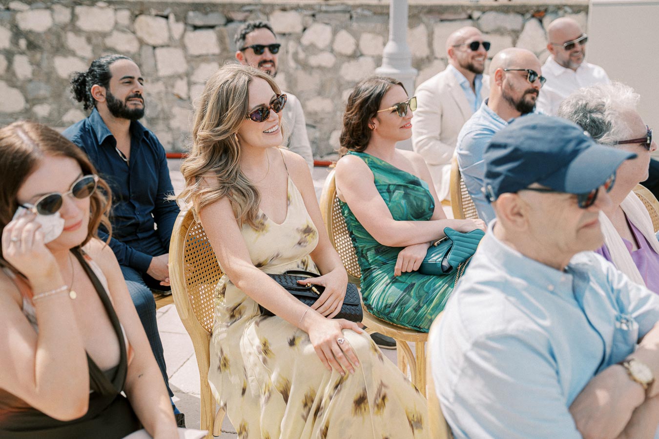 Group of people sitting outdoors at a sunny event, dressed in formal attire with sunglasses.