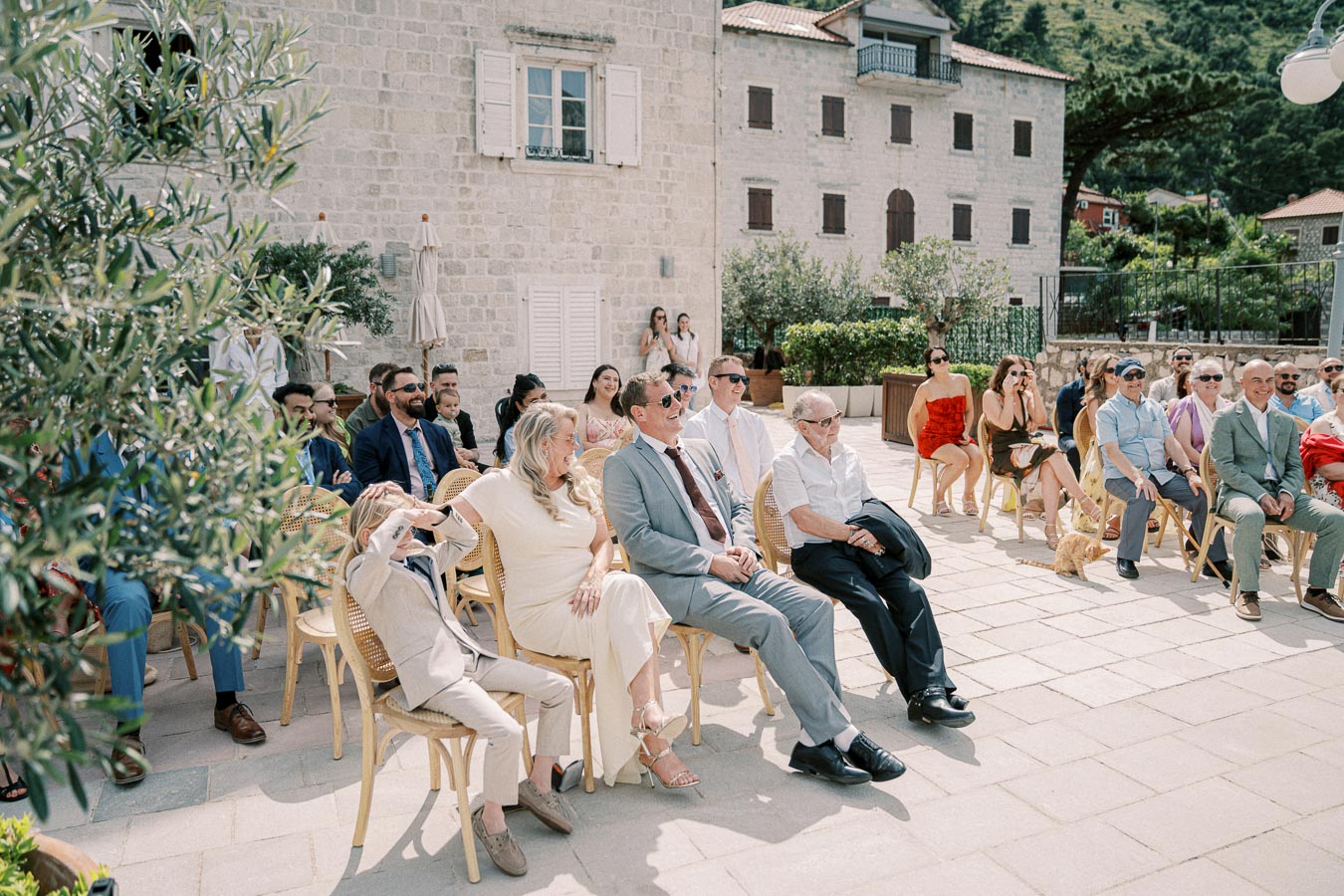 Outdoor wedding ceremony with seated guests in elegant attire, sunlit courtyard featuring stone buildings and greenery, festive summer celebration ambiance.