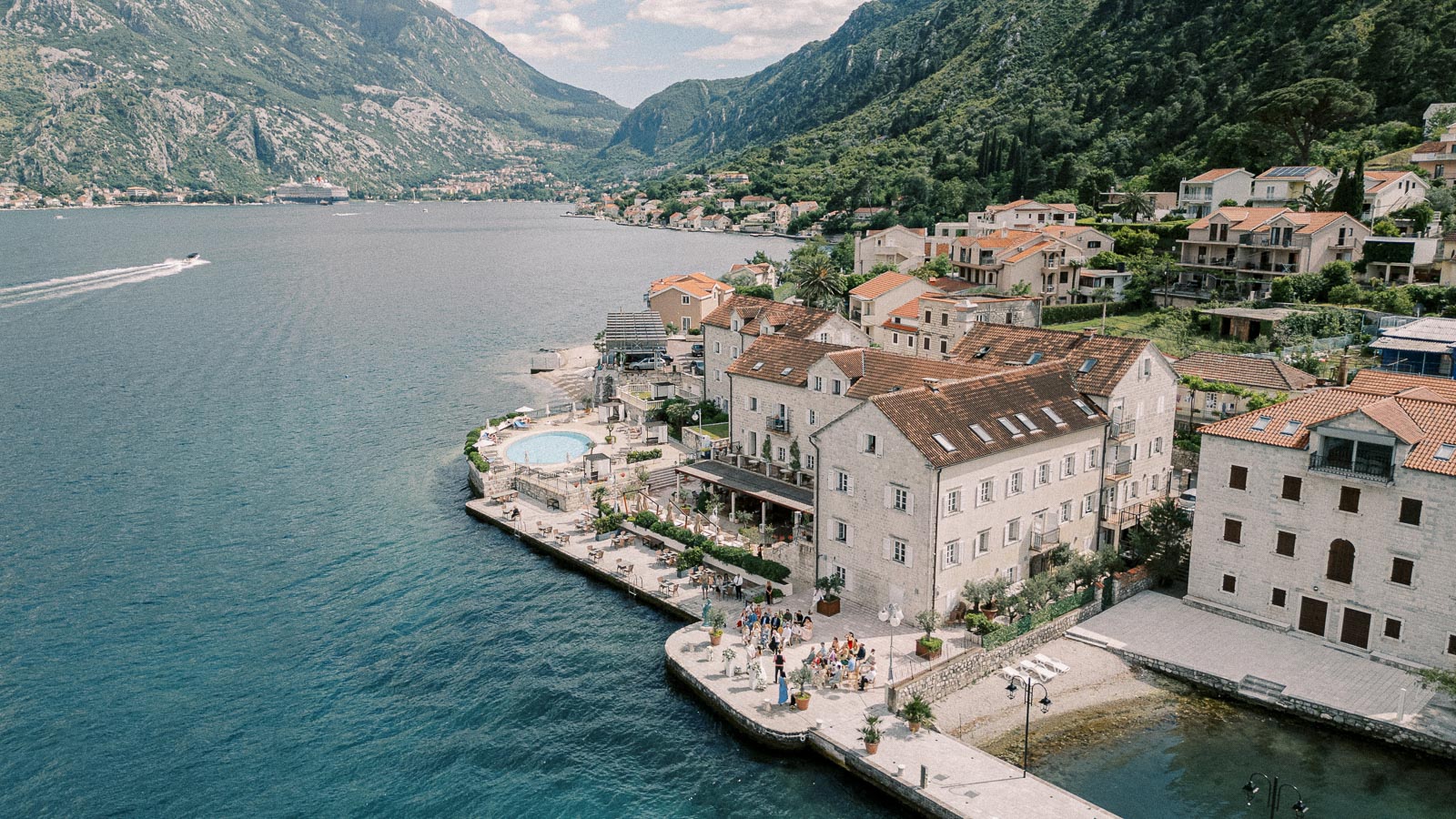 Aerial view of a picturesque coastal village with historic stone buildings and red-tiled roofs along the Adriatic Sea, surrounded by lush green mountains and clear blue water, featuring a waterfront promenade and a small harbor with boats.