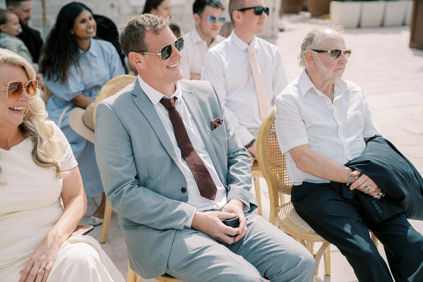 Group of well-dressed guests smiling and sitting outdoors at a sunny wedding ceremony, wearing sunglasses and formal attire.
