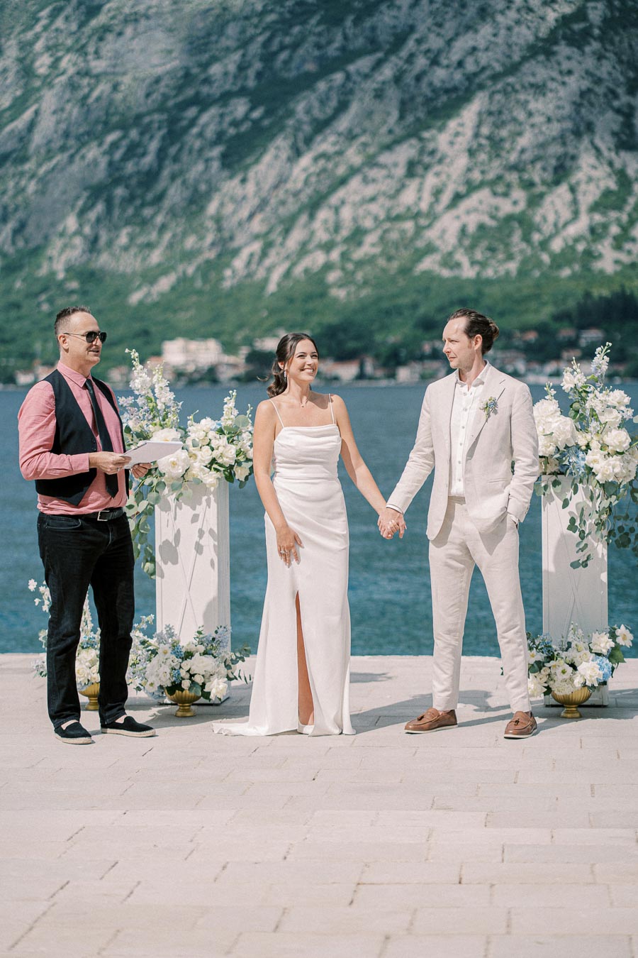 Wedding ceremony by the lake with a couple holding hands, officiant smiling, and a stunning mountainous backdrop.