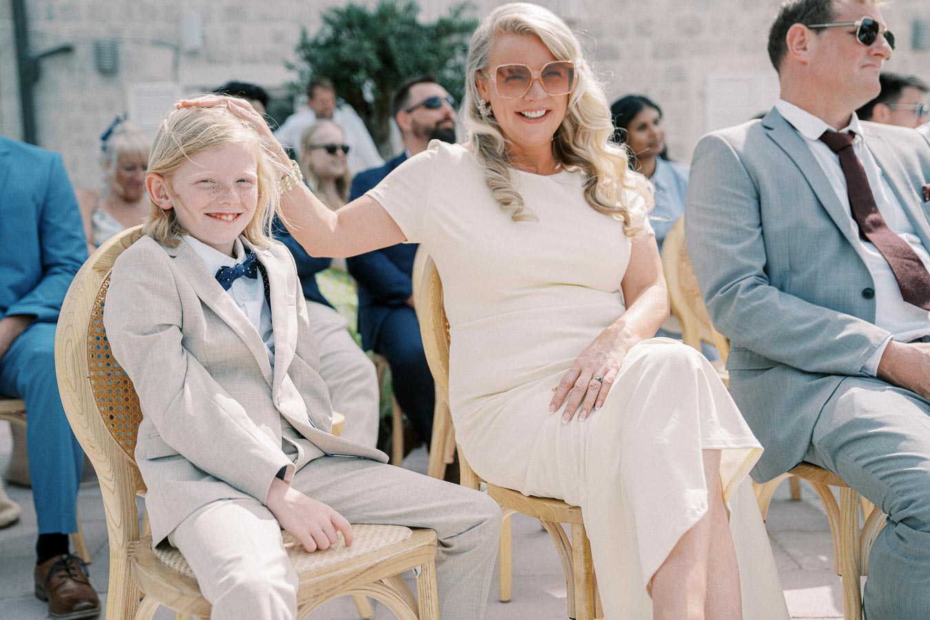 A smiling woman in sunglasses, wearing a light-colored dress, gently places her hand on a young boy's head. They are seated at an outdoor event, surrounded by other attendees in formal attire. The boy is dressed in a light suit with a bow tie, and both appear content in the sunny setting.