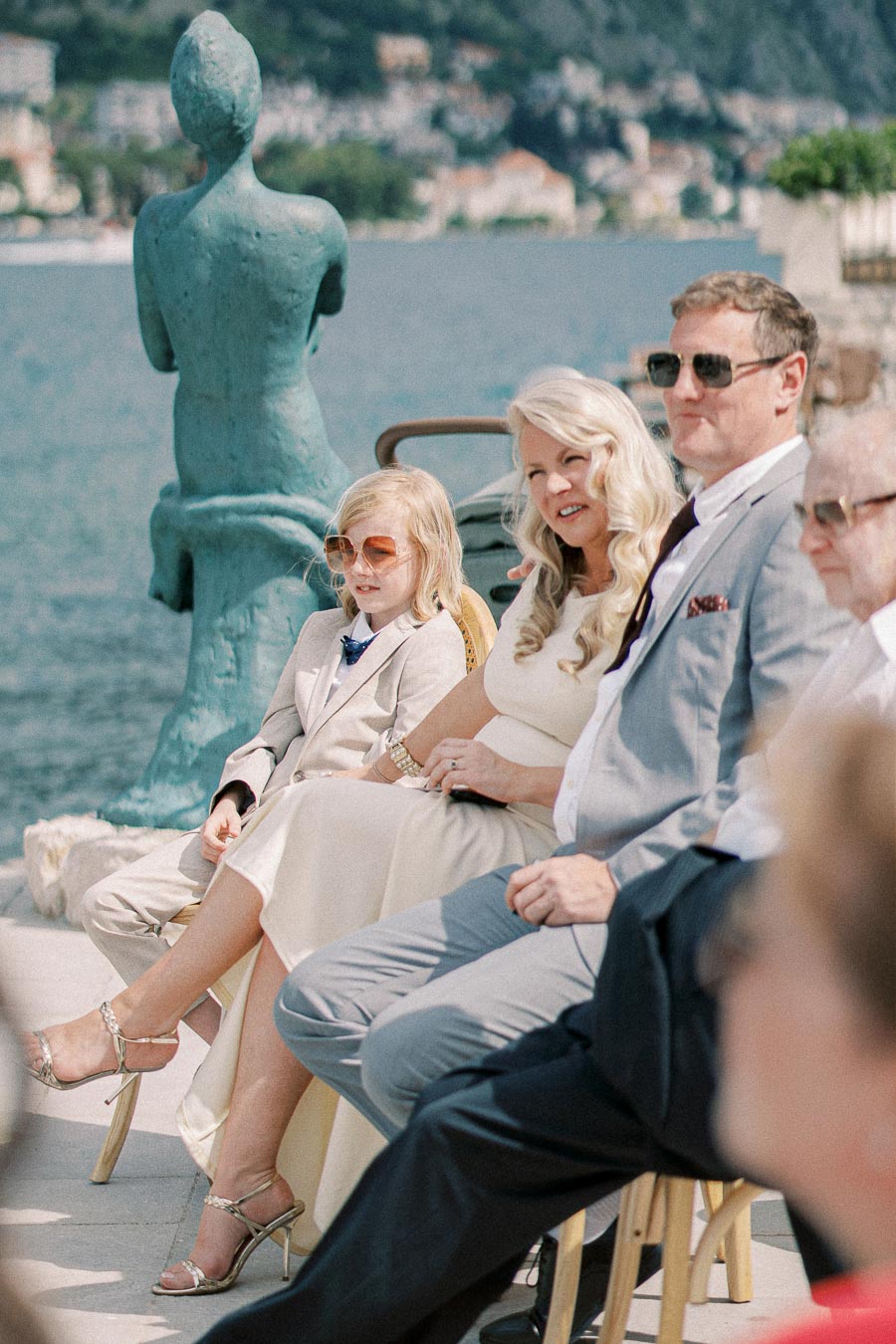 A group of people dressed elegantly sitting outdoors near a waterfront, with a statue in the background and scenic mountains across the water.