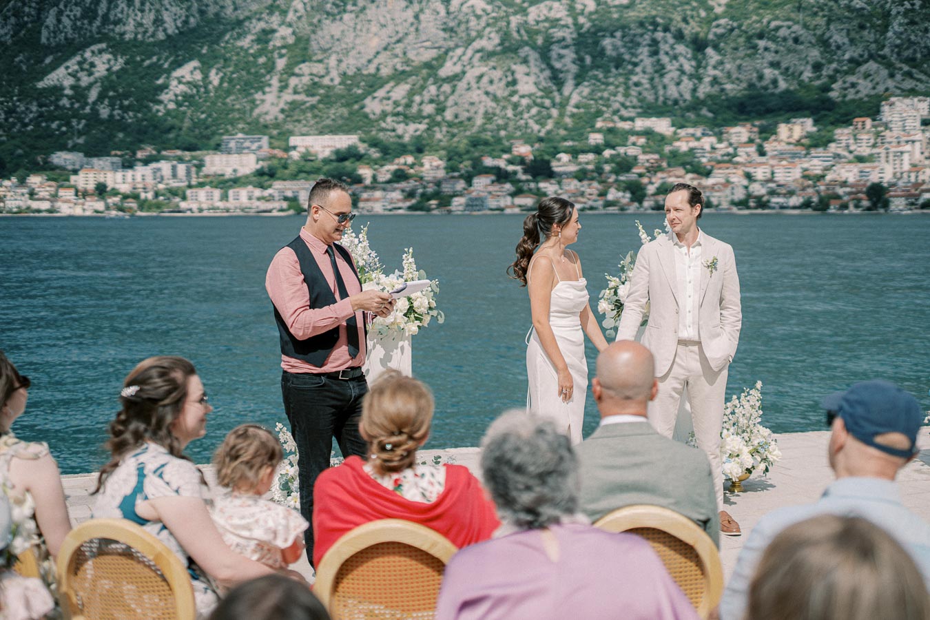 A lakeside wedding ceremony with a couple exchanging vows near a scenic mountain backdrop, surrounded by guests seated on wooden chairs.