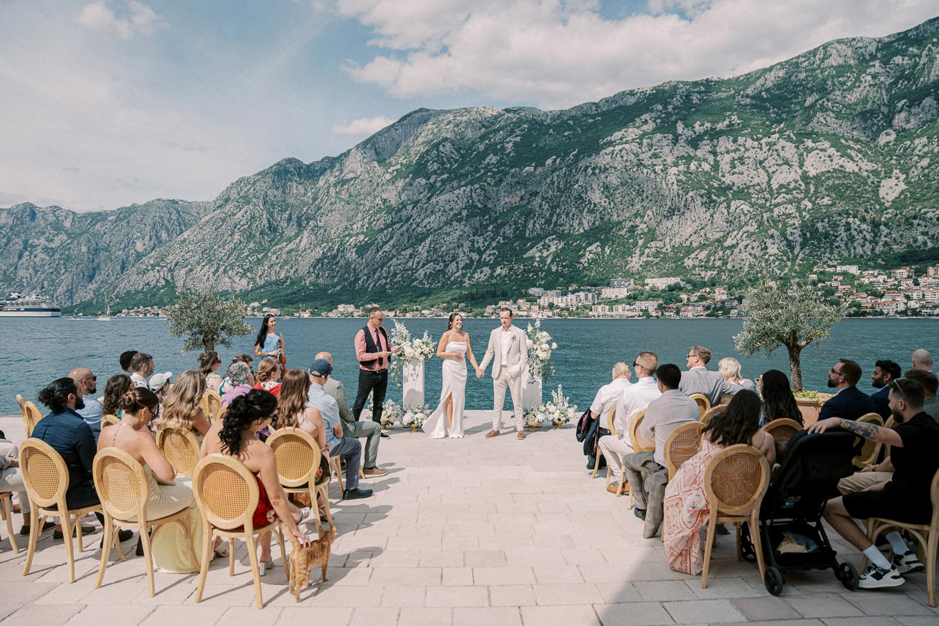 A scenic outdoor wedding ceremony by the sea, featuring a couple holding hands with mountainous backdrop, surrounded by elegantly dressed guests under a clear sky.