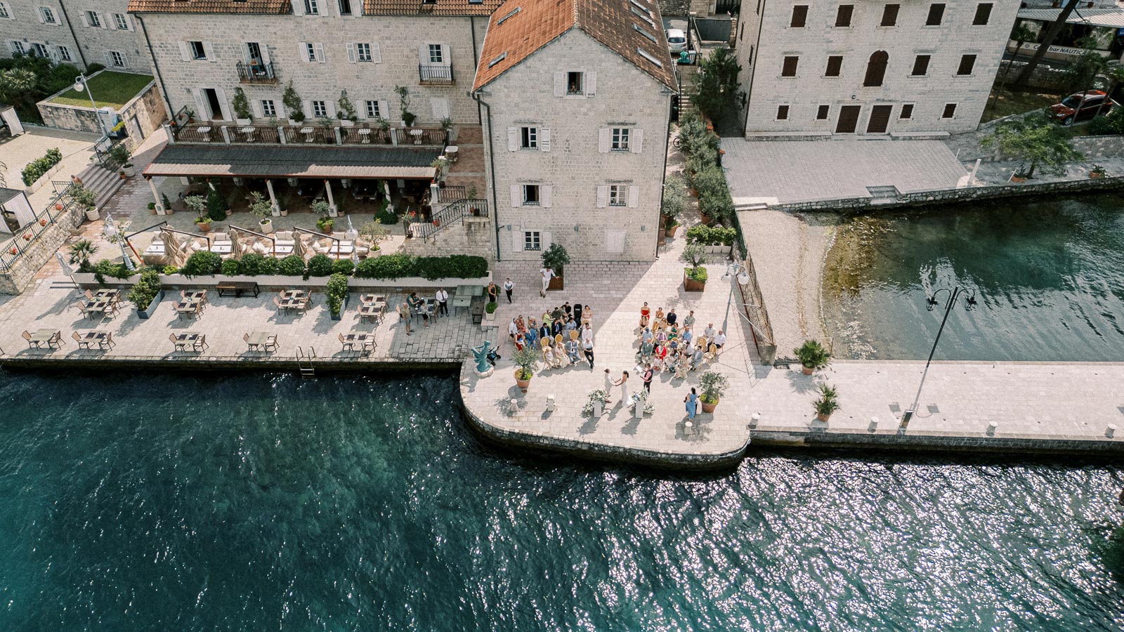 Aerial view of a picturesque waterfront wedding in a European coastal town, featuring a group of people gathered on a stone pier next to a historic building with outdoor seating by the water.