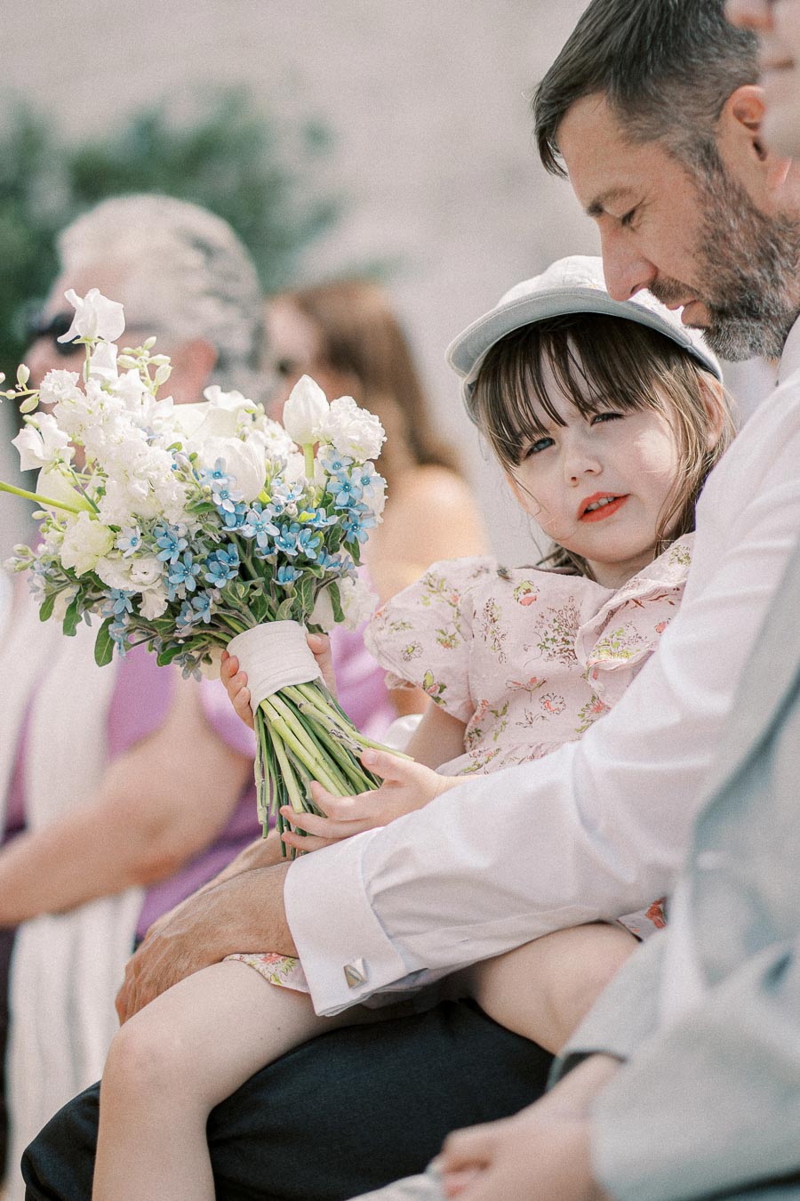 A young girl sits on a man's lap, holding a bouquet of white and blue flowers, while attending an outdoor event.