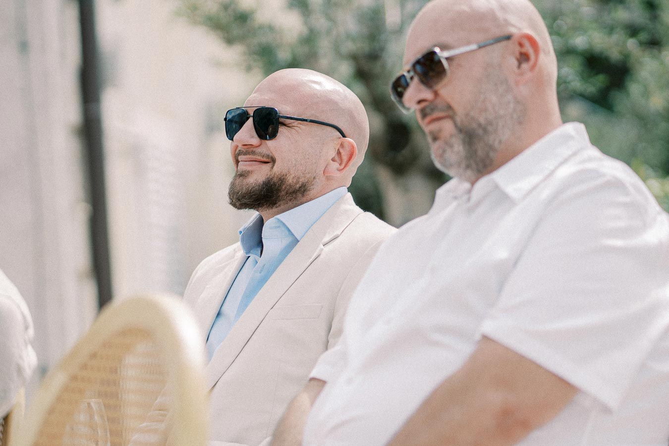 Two men wearing sunglasses attending an outdoor event, dressed in light-colored shirts with a blurred background of greenery.