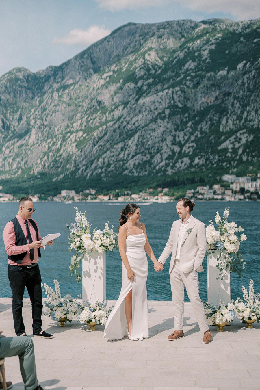 Elegant outdoor wedding ceremony by the sea, featuring a couple holding hands in front of a scenic mountain backdrop, with a celebrant officiating and floral arrangements enhancing the romantic atmosphere.