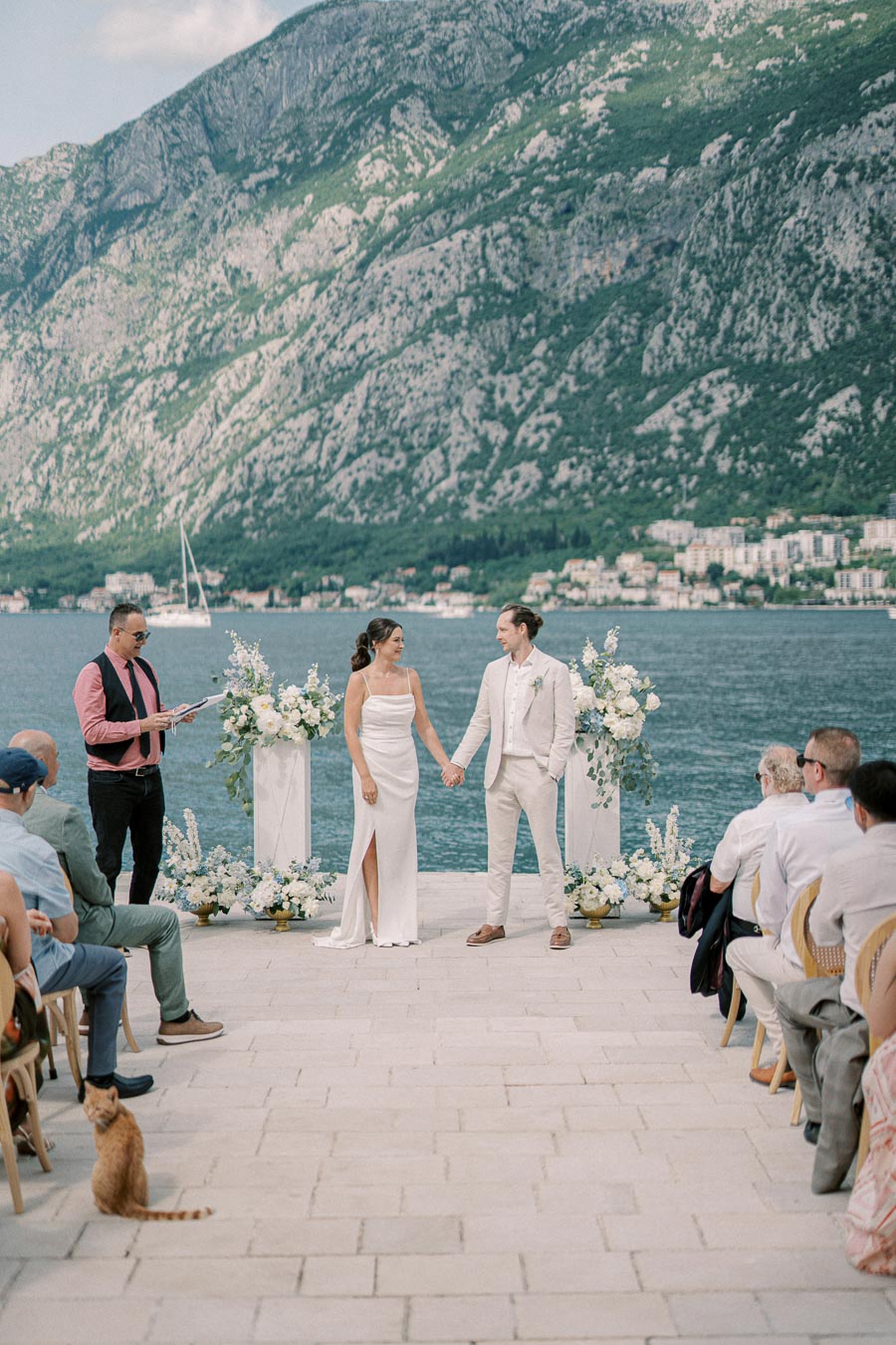 A bride and groom hold hands during a waterfront wedding ceremony, surrounded by floral arrangements, with a scenic mountain and sea backdrop.