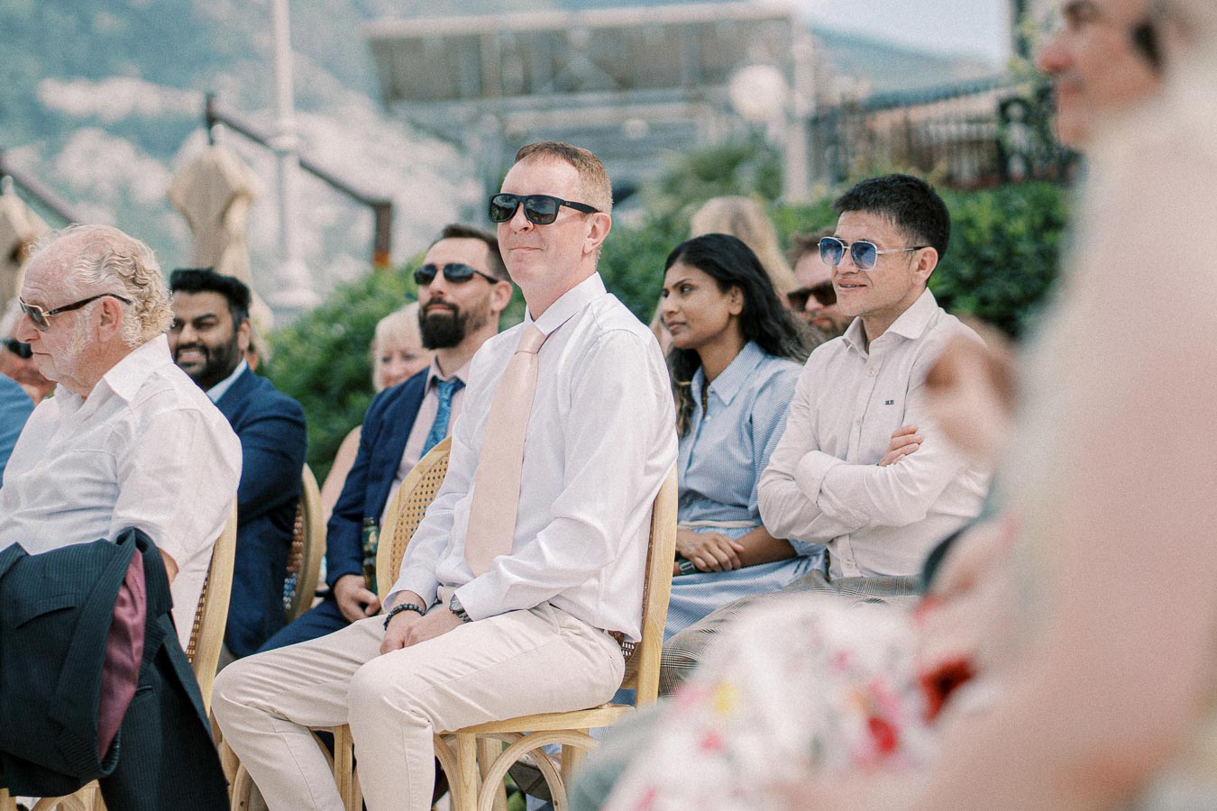 A group of people in formal attire sitting outdoors at a daytime event, with greenery and mountains in the background, conveying a serene and professional atmosphere.