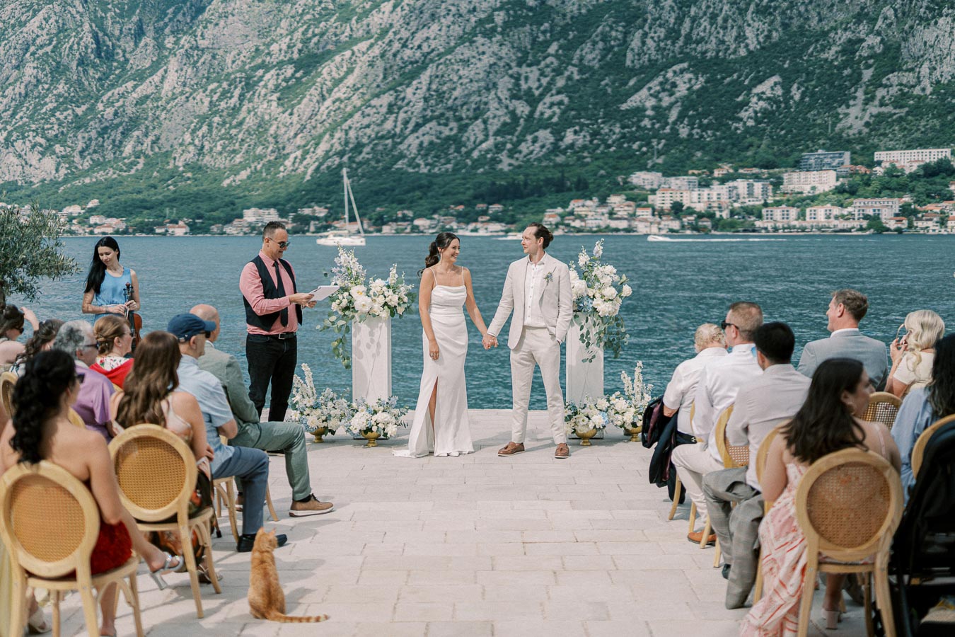 Scenic outdoor wedding ceremony by the water, with bride and groom holding hands, surrounded by elegantly dressed guests and floral arrangements, cliffs and buildings in the background.