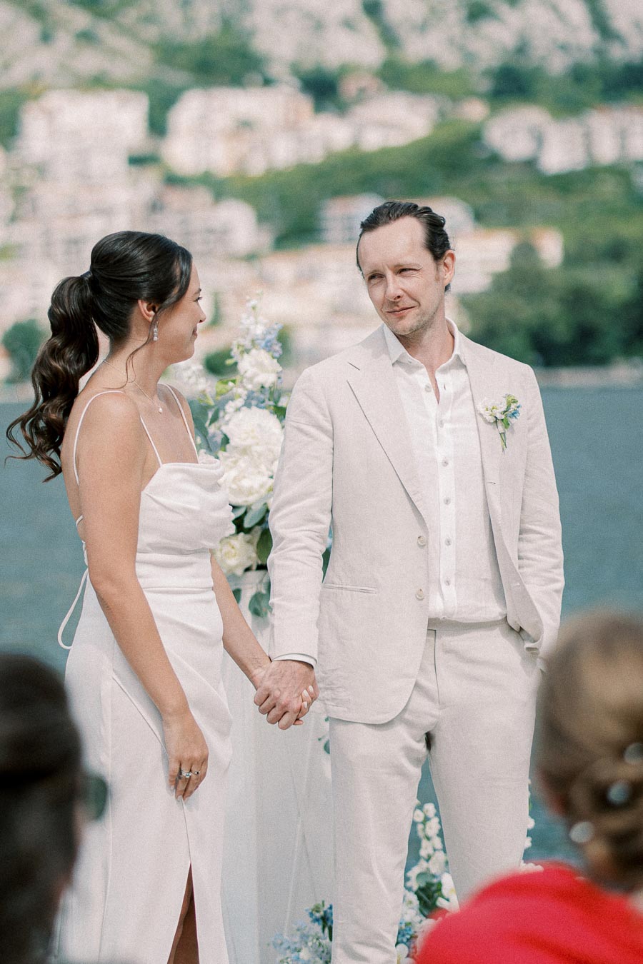 Elegant couple holding hands during an outdoor wedding ceremony by the water, with lush greenery and buildings in the background.