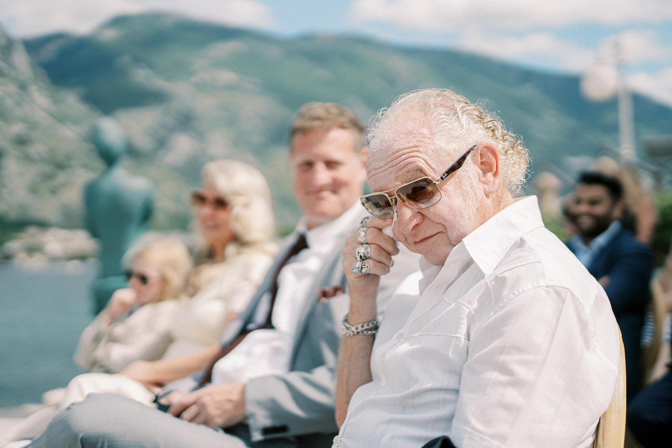 Elderly man in sunglasses and white shirt sitting outdoors at an event with blurred people in the background, with scenic mountains and water view.