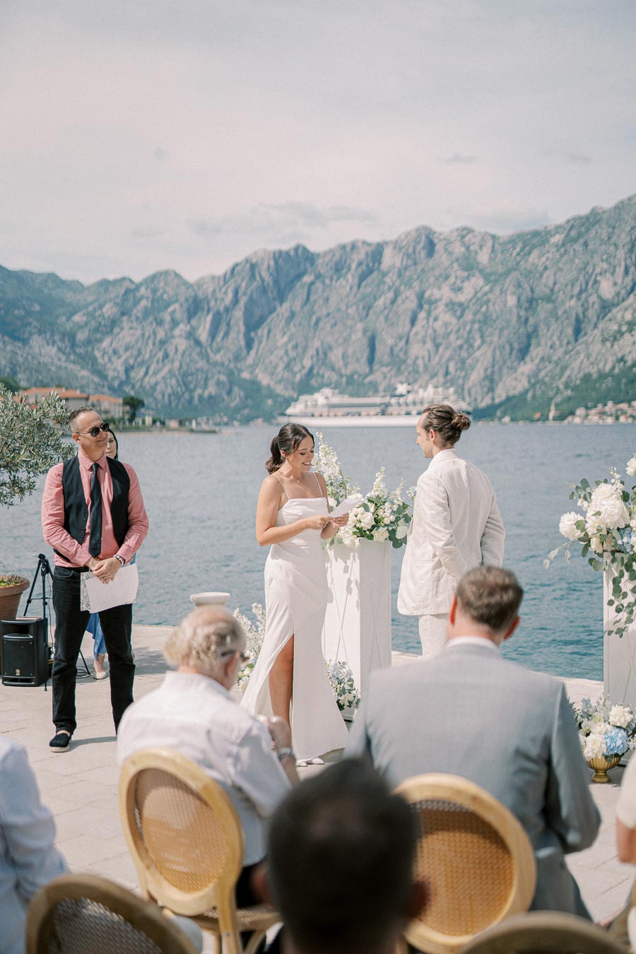 Elegant outdoor wedding ceremony by the sea with a mountainous backdrop, featuring a bride in a white dress holding vows and a groom in a light suit, surrounded by guests and floral arrangements.