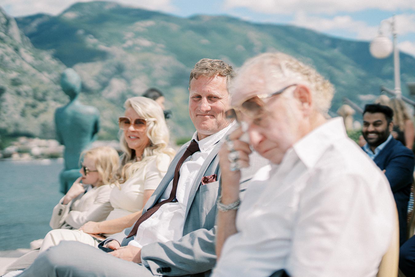 Group of people sitting outdoors with a scenic mountain and lake view in the background, dressed in formal attire and enjoying a sunny day.