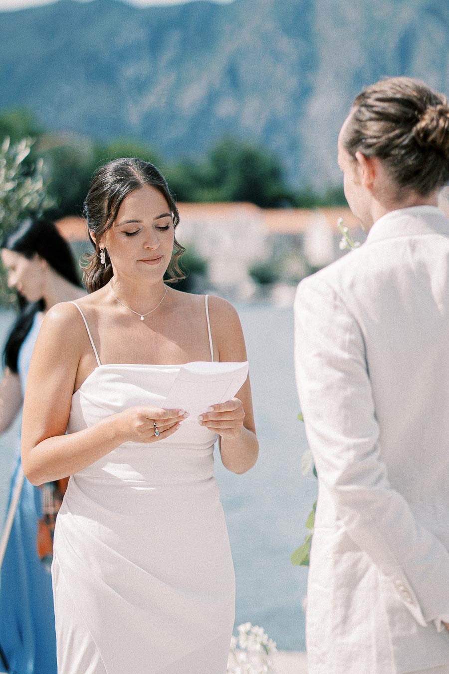 Bride reading vows during outdoor wedding ceremony by the water, with mountains in the background.