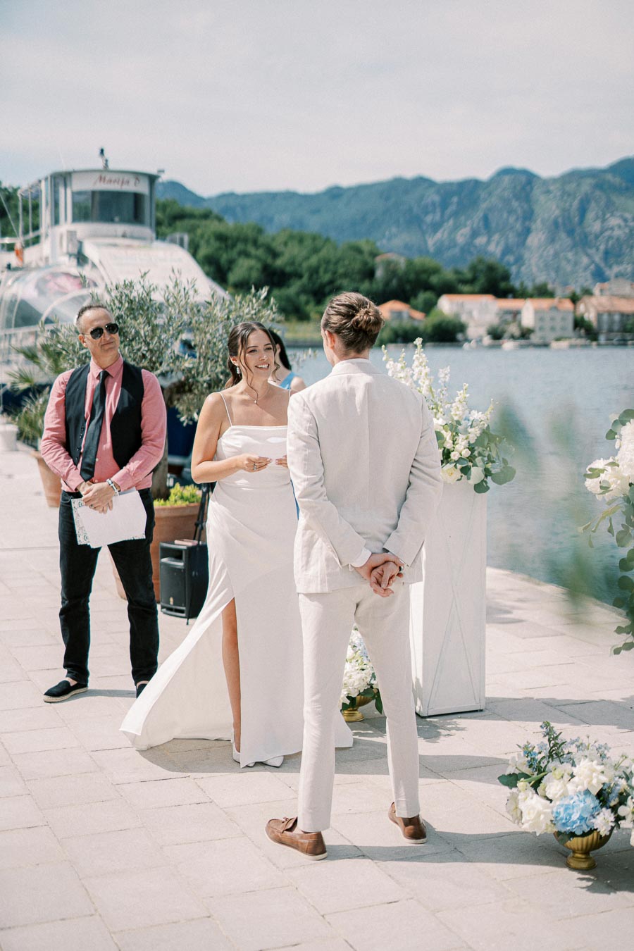 A couple exchanging vows during an outdoor wedding ceremony by a scenic lakeside with mountains in the background. A man stands beside them holding a script. The bride is in a white dress, and the groom wears a light-colored suit. The setting includes floral decorations and a boat visible on the water.