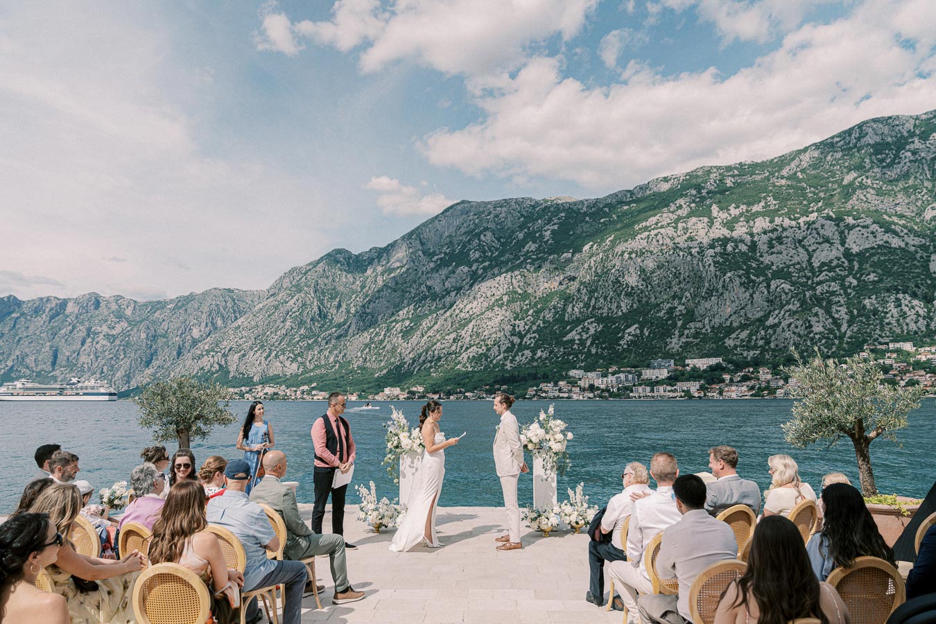 Outdoor wedding ceremony by a scenic lake with mountains in the background, attended by seated guests, focusing on the couple exchanging vows under a partly cloudy sky.