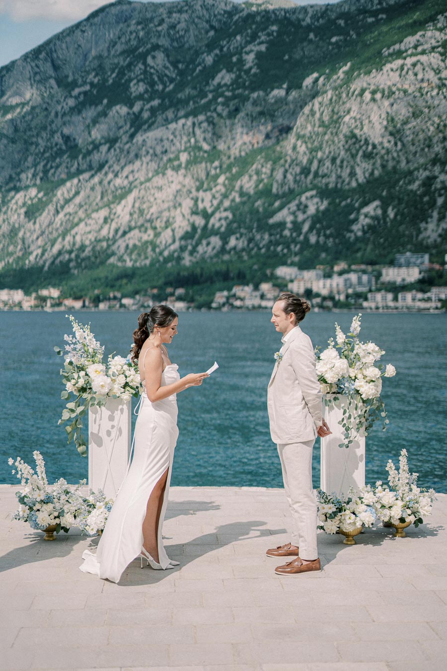 Wedding ceremony by the sea with bride and groom exchanging vows; mountainous landscape and floral arrangements in the background.