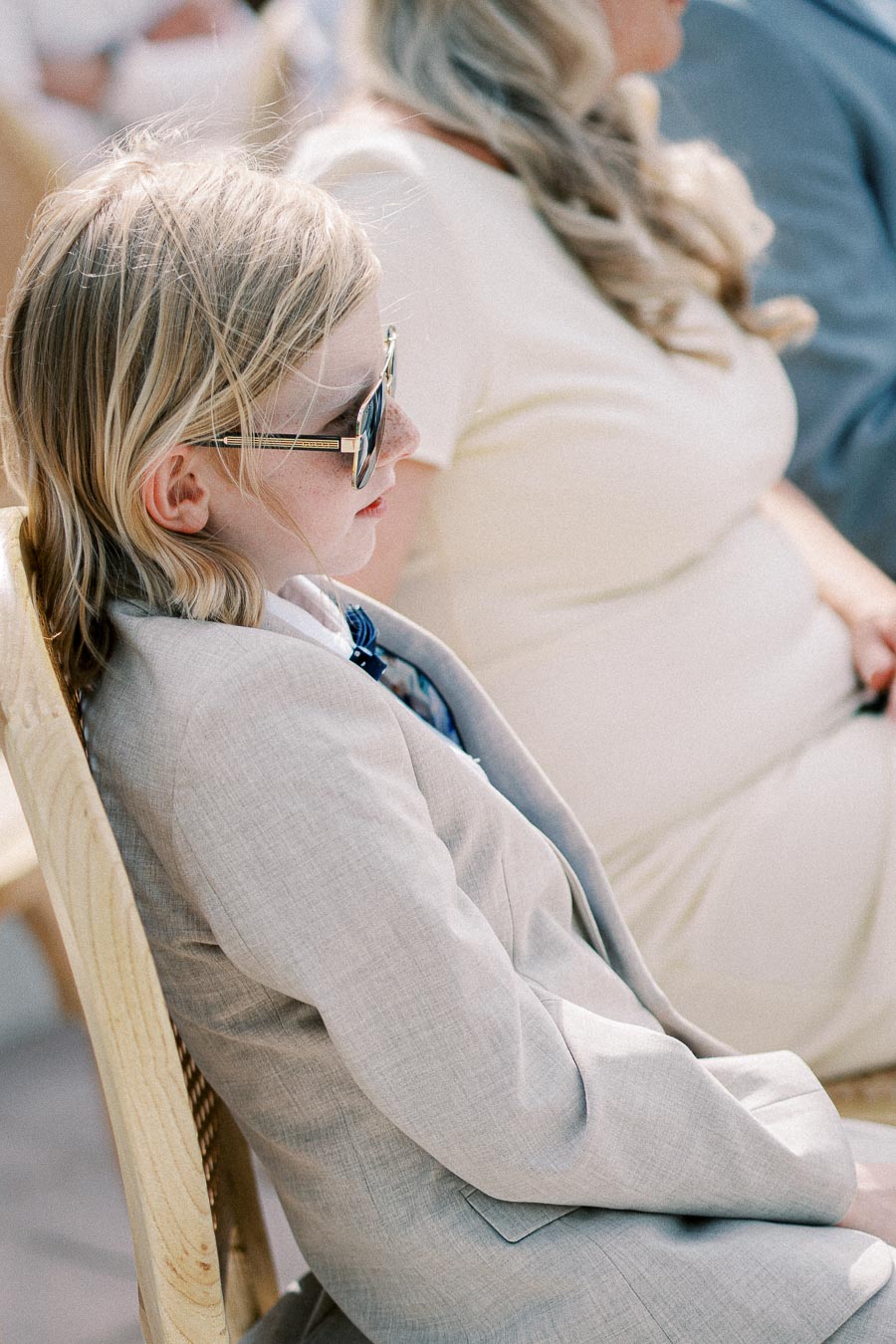A young child wearing sunglasses and a light gray suit sits attentively on a wooden chair during an outdoor event.