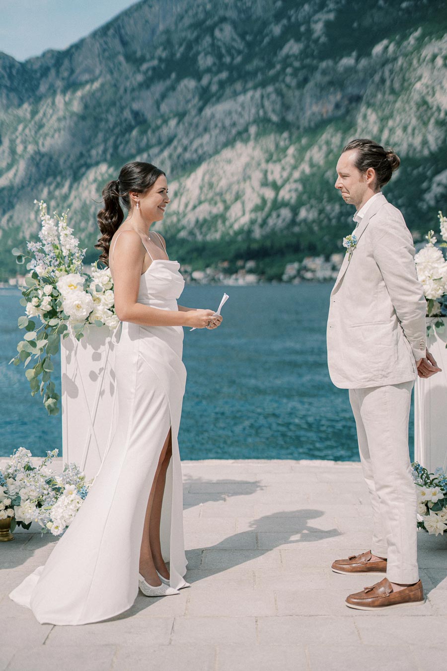 A bride and groom exchange vows during a picturesque outdoor wedding ceremony by the water, with mountains in the background. She wears a white gown, while he wears a light suit, surrounded by floral arrangements.