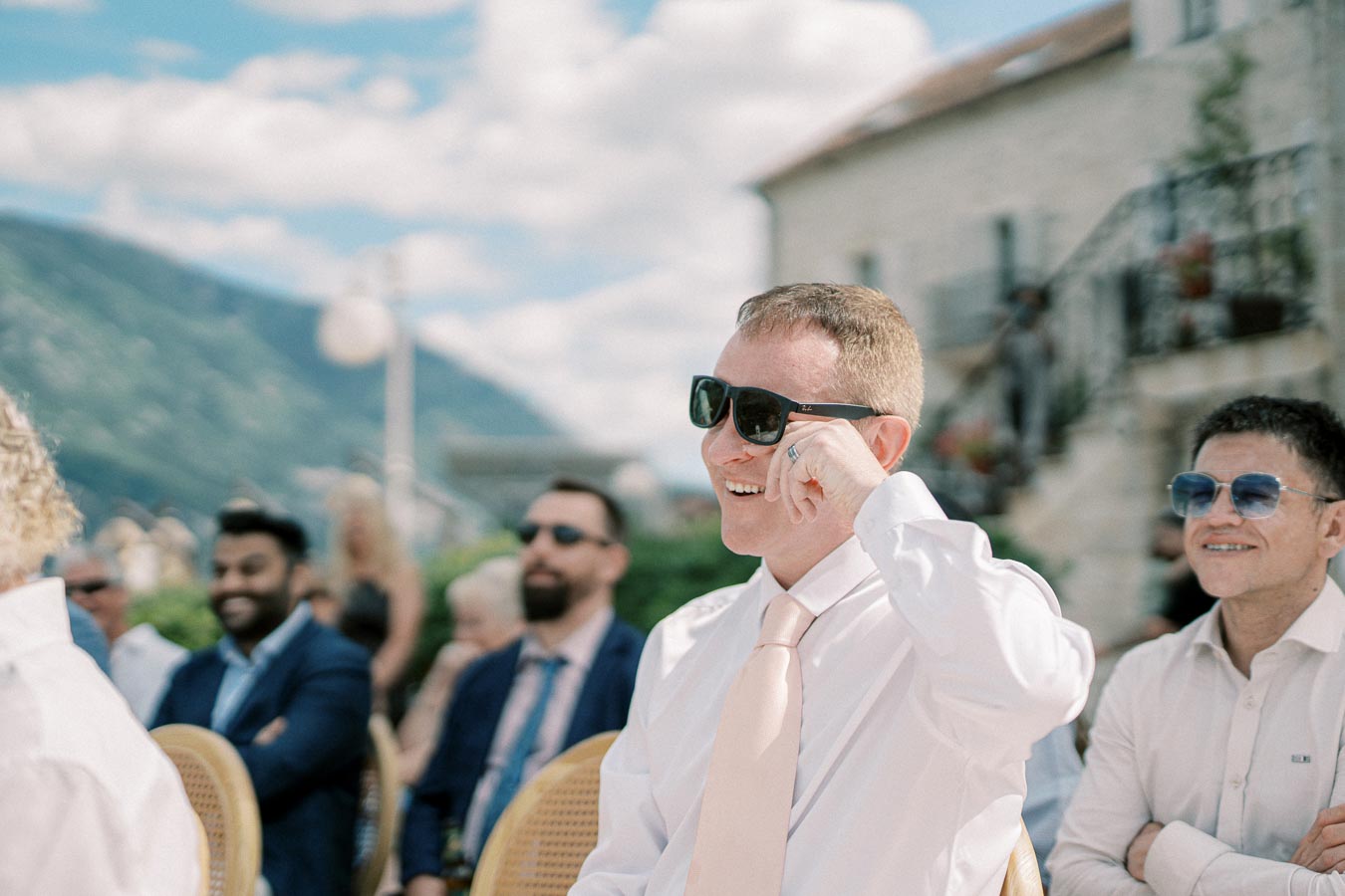 A joyful man wearing sunglasses and a white shirt with a pink tie sits among guests at an outdoor event, set against a backdrop of mountains and a partially cloudy sky.